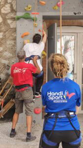 Deux moniteurs accompagnent une femme lors d'une séance d'escalade en salle. La grimpeuse est en train d'attraper une prise.