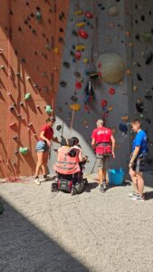 Trois accompagnateurs et une personne en fauteuil se tiennent de dos, face à un mur d'escalade. La personne en fauteuil s'apprête à grimper le mur.