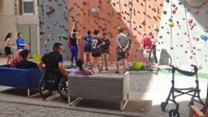 Un groupe de personnes est réuni au pied d'un mur d'escalade en intérieur. Une personne est en fauteuil roulant, un déambulateur est visible sur la droite de la photo. Des moniteurs sont en train d'équiper les grimpeurs qui s'apprêtent à escalader le mur.