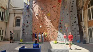 Un groupe de personnes sont dos à la photo lors d'une séance d'escalade sur mur, en intérieur. Les grimpeurs sont tous accompagnés par un moniteur au sol.