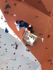 Un grimpeur est au centre de la photo, en train d'escalader un dévers. Le grimpeur a une prothèse à la jambe gauche.