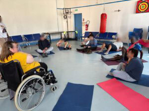 Groupe de personnes assises en cercle sur des tapis de relaxation. Une femme participe également à l'activité sur un fauteuil roulant
